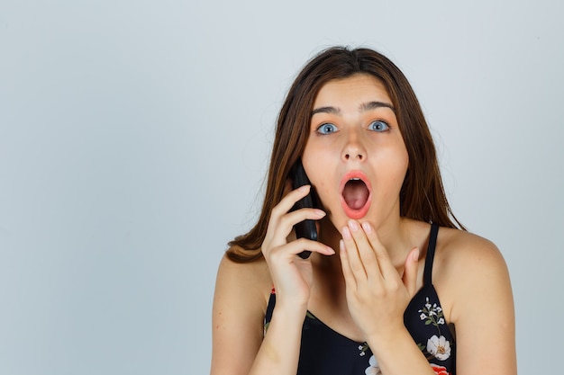 Young lady in blouse talking on smartphone, holding hand near open mouth and looking surprised , front view.