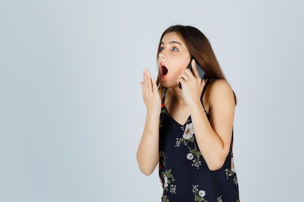 Giovane donna in camicetta che parla su smartphone, tenendo la mano vicino alla bocca aperta e guardando sorpreso, vista frontale.
