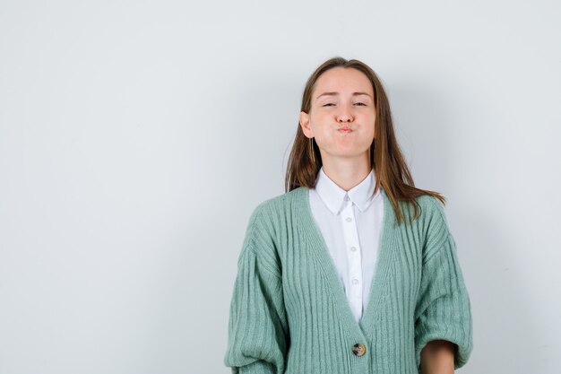 Young lady blowing cheeks while looking at front in shirt, cardigan and looking funny , front view.