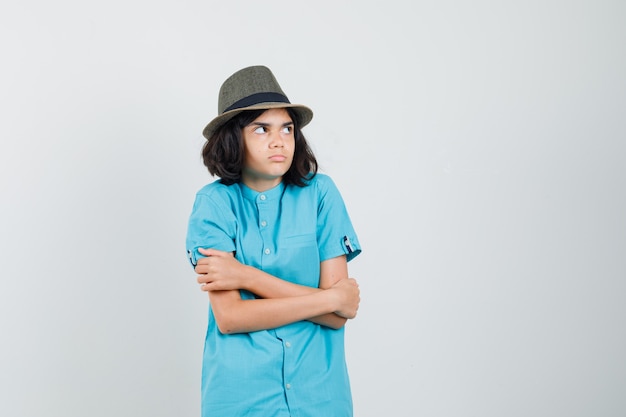 Young lady in blue shirt, hat hugging herself and looking uncomfortable