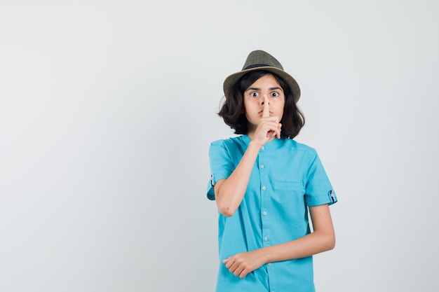 Young lady in blue shirt, hat showing silence gesture and looking alarmed