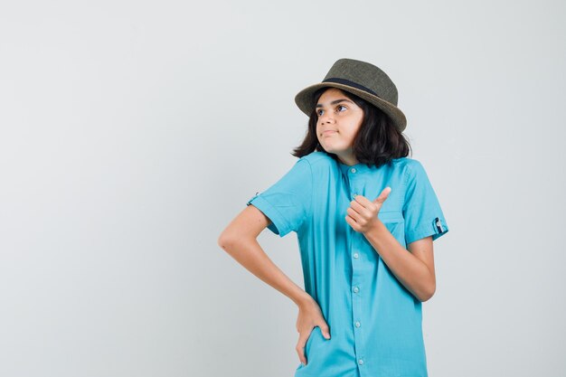 Молодая дама в синей рубашке, шляпа показывает палец вверх и выглядит довольной