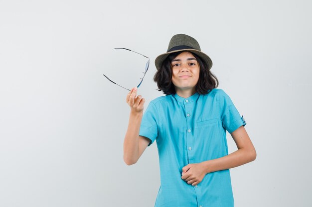 Young lady in blue shirt, hat, sunglasses showing helpless gesture while holding glasses