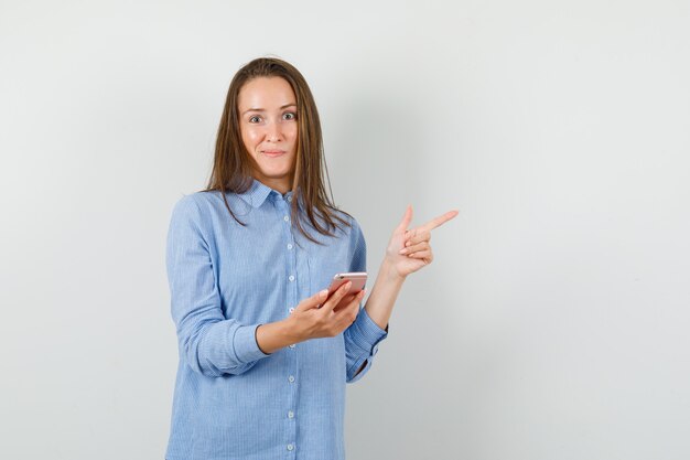 Young lady in blue shirt holding mobile phone while pointing away and looking curious