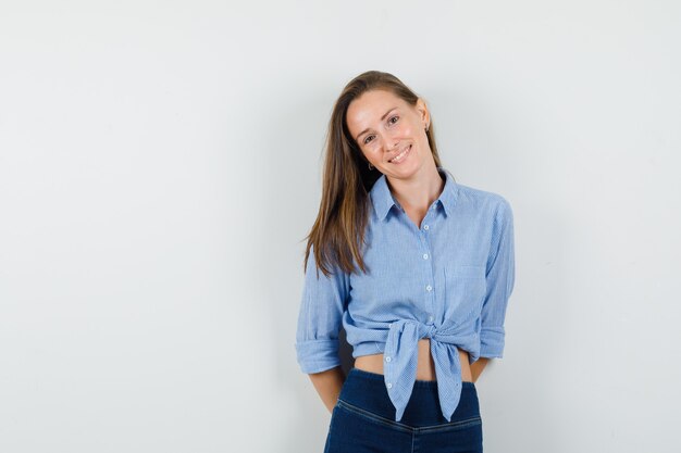 Giovane donna in camicia blu, pantaloni tenendosi per mano sulla schiena e guardando ottimista