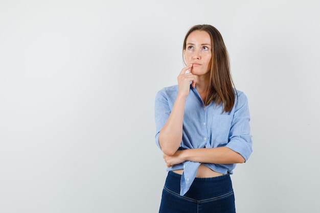 Young lady in blue shirt, pants looking up with finger near mouth and looking pensive