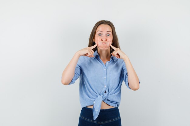 Young lady in blue shirt, pants pressing on blowing cheeks and looking funny