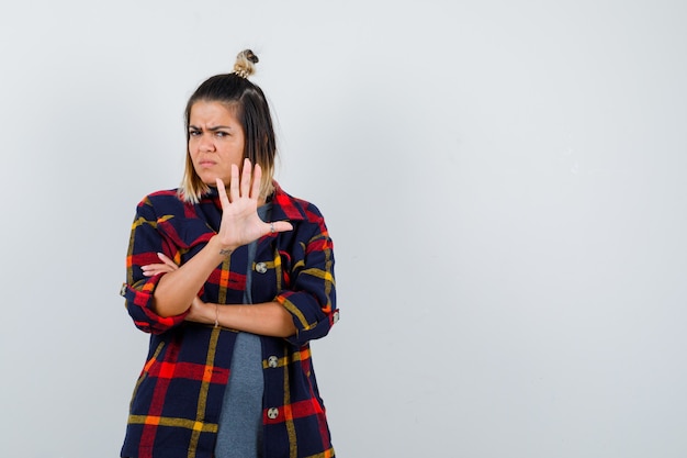 Young lady in casual checked shirt showing stop gesture and looking dismal , front view.