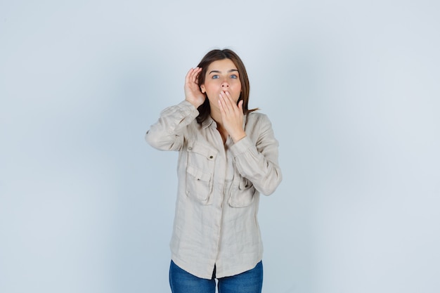 Young lady in casual, jeans keeping one hand behind ear and the other on mouth and looking shocked , front view.