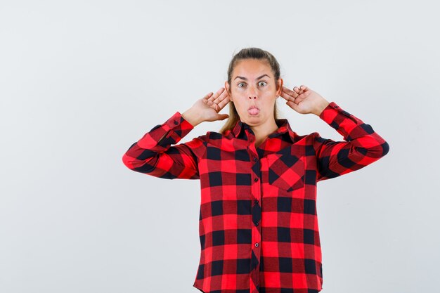 Young lady in casual shirt holding fingers behind ears and looking funny , front view.