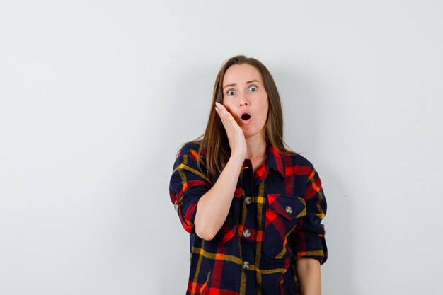 Young lady in casual shirt keeping hand on cheek and looking surprised , front view.