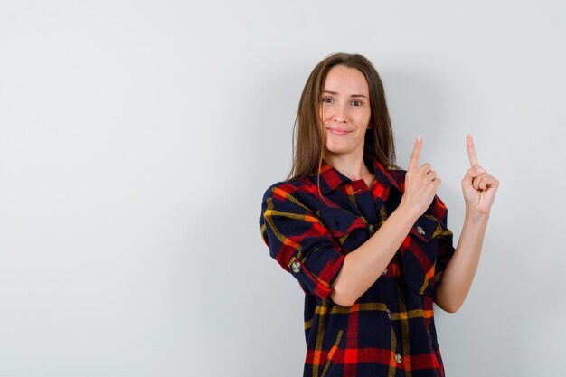 Young lady in casual shirt pointing up and looking confident , front view.