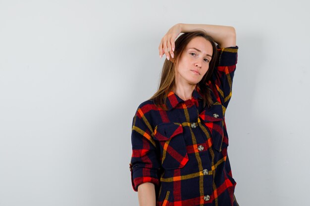 Young lady in casual shirt posing with raised arm on head and looking elegant , front view.