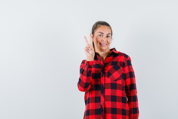 Young lady in casual shirt showing V-sign and looking merry , front view.