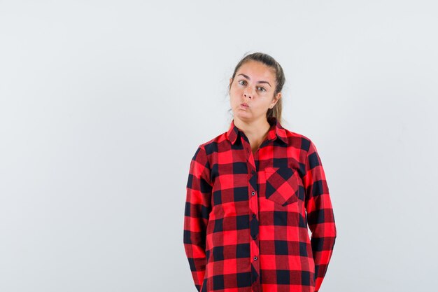 Young lady in checked shirt blowing cheeks and looking hesitant