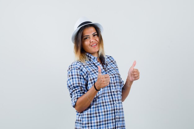 Young lady in checked shirt hat showing double thumbs up and looking confident  