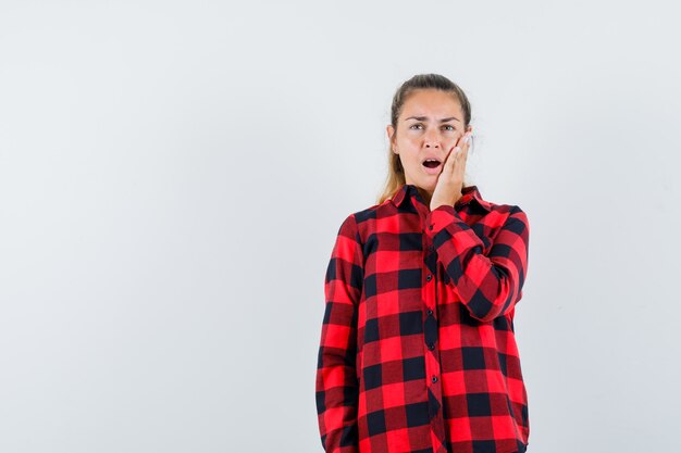 Young lady in checked shirt holding hand on cheek and looking forgetful