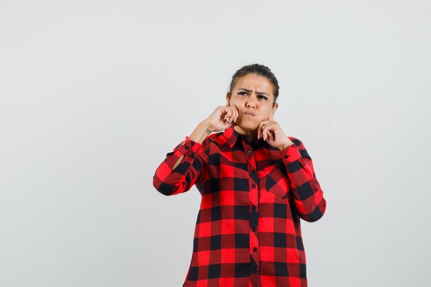 Young lady in checked shirt holding hands on blowing cheeks and looking pensive