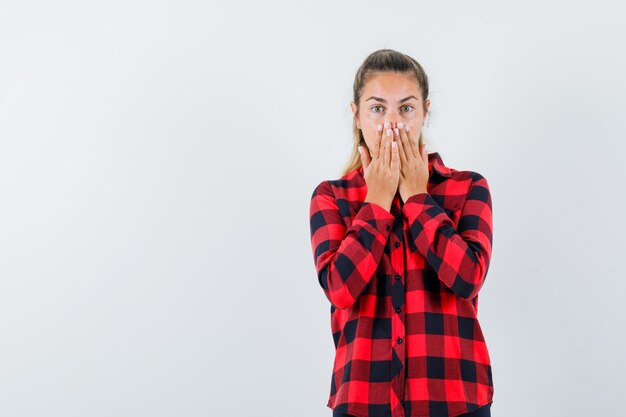 Young lady in checked shirt holding hands on mouth and looking surprised