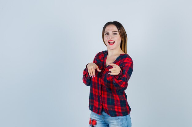 Young lady in checked shirt, jeans pointing at front while opening mouth and looking happy , front view.