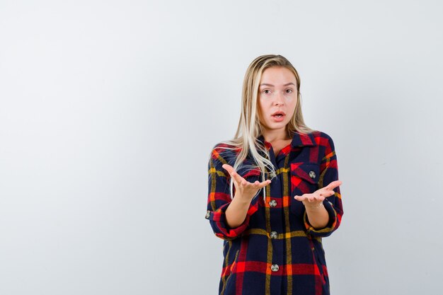 Young lady in checked shirt keeping hands in aggressive manner and looking puzzled , front view.