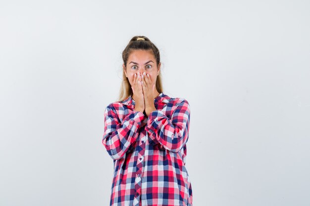 Young lady in checked shirt keeping hands on mouth and looking scared , front view.