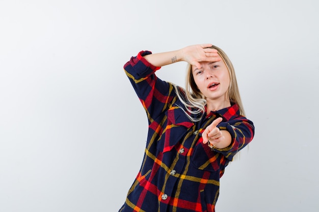 Young lady in checked shirt pointing aside while holding hand on forehead and looking positive , front view.