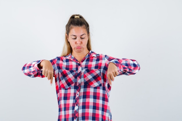 Free photo young lady in checked shirt pointing down, curving lips and looking focused , front view.