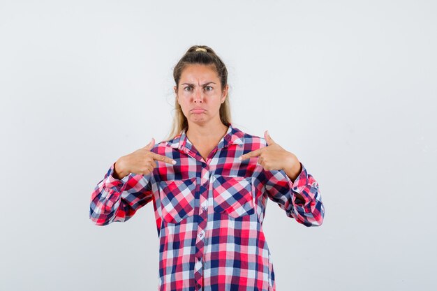 Young lady in checked shirt pointing at herself and looking serious , front view.