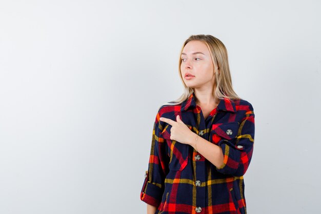 Young lady in checked shirt pointing to the left side and looking confident , front view.