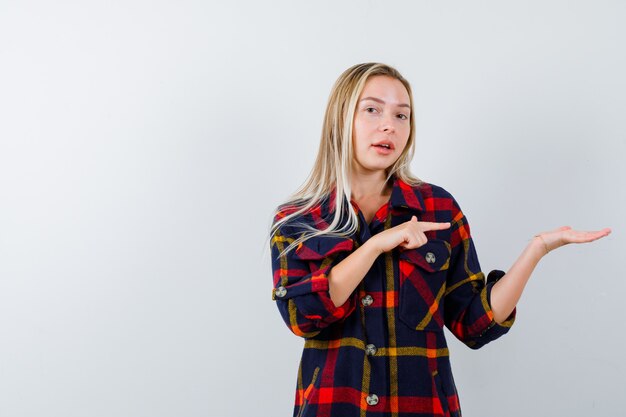 Young lady in checked shirt pointing at palm and looking confident , front view.