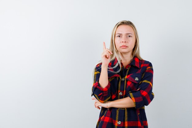 Young lady in checked shirt pointing up and looking confident , front view.