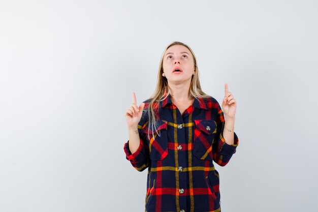 Young lady in checked shirt pointing up and looking puzzled , front view.