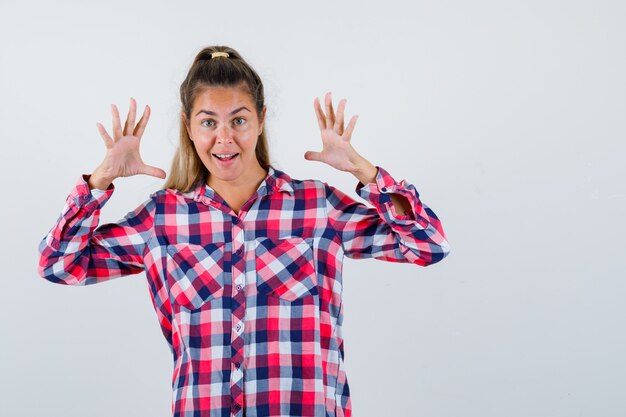 Young lady in checked shirt raising hands and looking happy , front view.