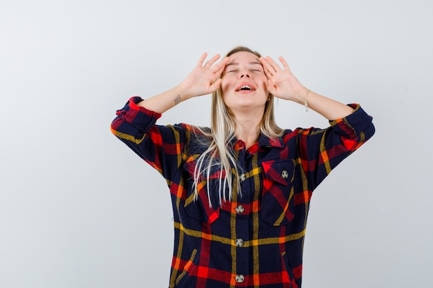 Foto gratuita giovane donna in camicia a quadri che mostra le palme sul viso e sembra pacifica, vista frontale.