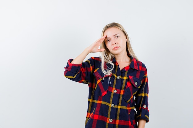 Giovane donna in camicia a quadri che mostra gesto di saluto e guardando fiducioso, vista frontale.