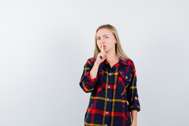 Giovane donna in camicia a quadri che mostra gesto di silenzio e che sembra serio, vista frontale.