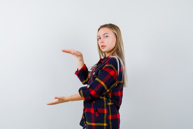 Young lady in checked shirt showing size sign while looking over shoulder and looking confident , front view.