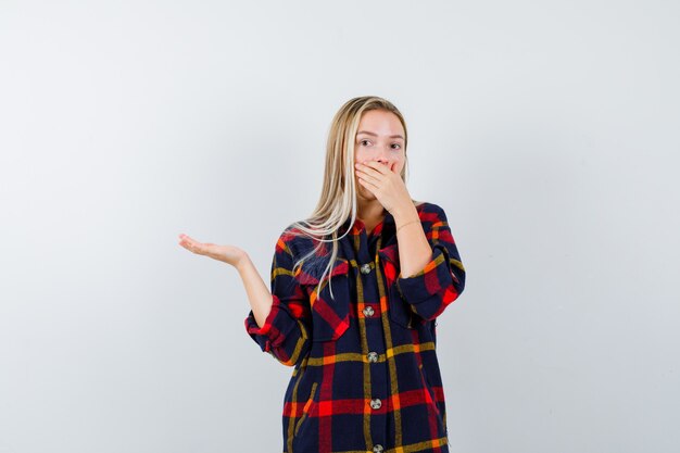 Young lady in checked shirt showing something while holding hand on mouth and looking amazed , front view.