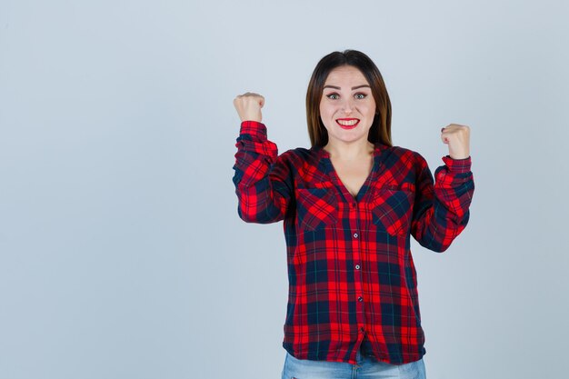 Young lady in checked shirt showing winner gesture and looking glad , front view.