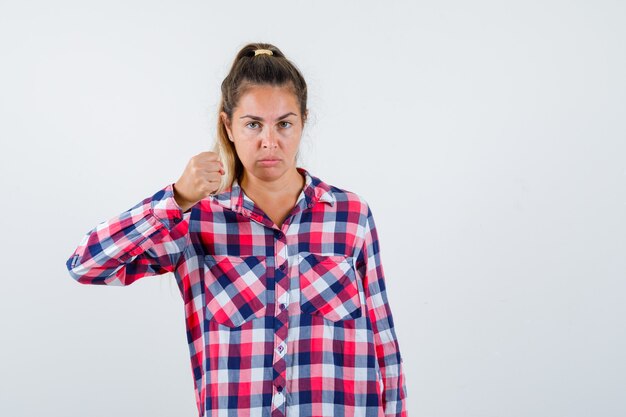 Young lady in checked shirt threatening with fist and looking strict , front view.