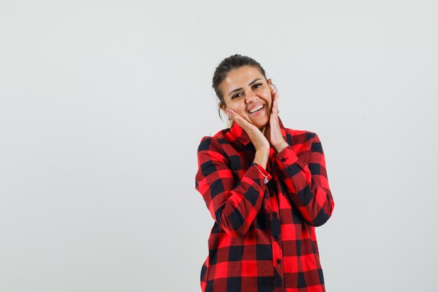 Young lady in checked shirt touching face skin on cheeks and looking delicate