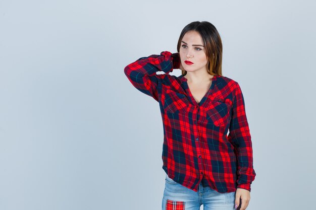 Young lady in checked shirt with hand behind neck while looking away and looking thoughtful , front view.