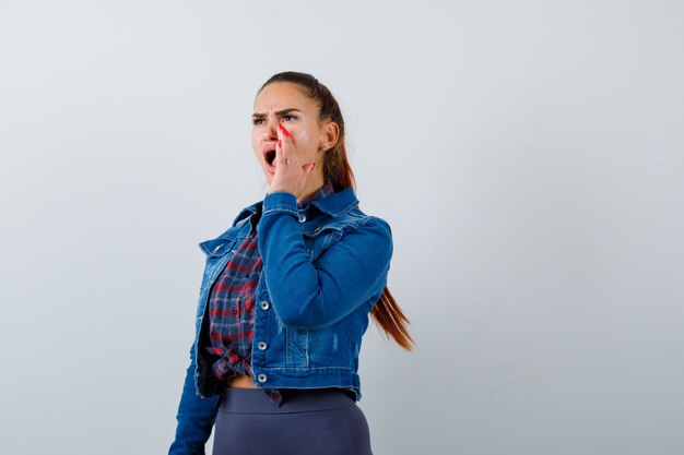 Young lady in checkered shirt, denim jacket shouting while with hand near mouth and looking annoyed , front view.