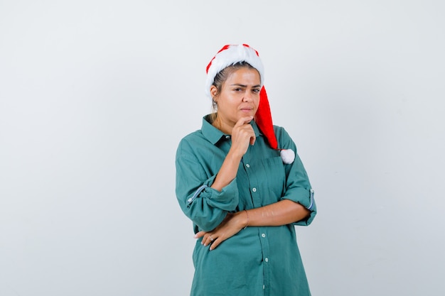 Giovane donna con cappello di natale, camicia con il dito sul mento e sguardo pensieroso, vista frontale.