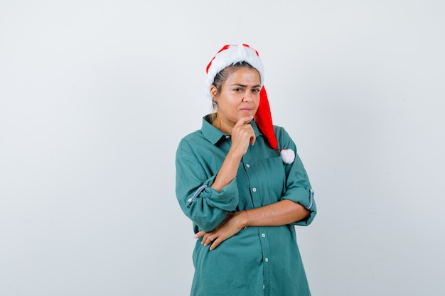 크리스마스 모자를 쓴 젊은 여성, 턱에 손가락을 대고 생각에 잠긴 모습을 한 셔츠, 전면 보기.