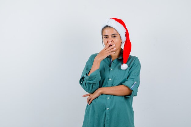 Young lady in christmas hat, shirt with hand on mouth and looking perplexed , front view.