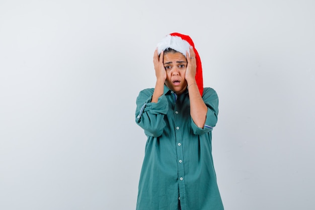 Giovane donna con cappello di natale, camicia con le mani sulla testa e sguardo spaventato, vista frontale.