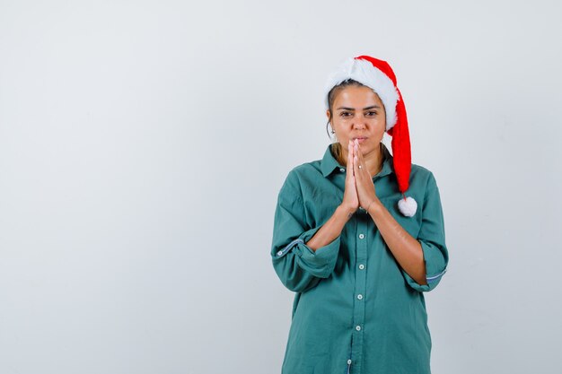 Young lady in christmas hat, shirt with hands in praying gesture and looking hopeful , front view.