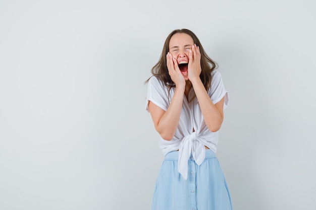 Young lady clasping face with hands while screaming in white blouse,blue skirt and looking joyous